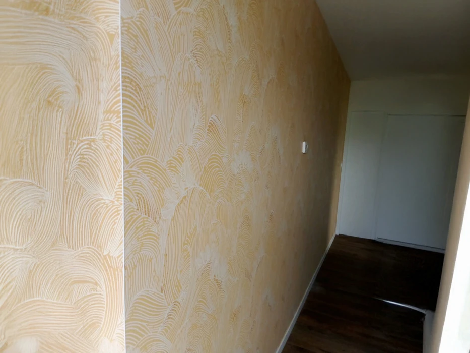 Intérieur résidentiel avec mur d'accent décoratif présentant un enduit texturé beige aux motifs ondulés organiques. Application artisanale d'un revêtement mural à effet relief, technique de finition décorative au peigne ou spatule crantée créant des vagues fluides. Rendu mat uniforme sur support préparé, démonstration de savoir-faire en décoration murale haut de gamme.