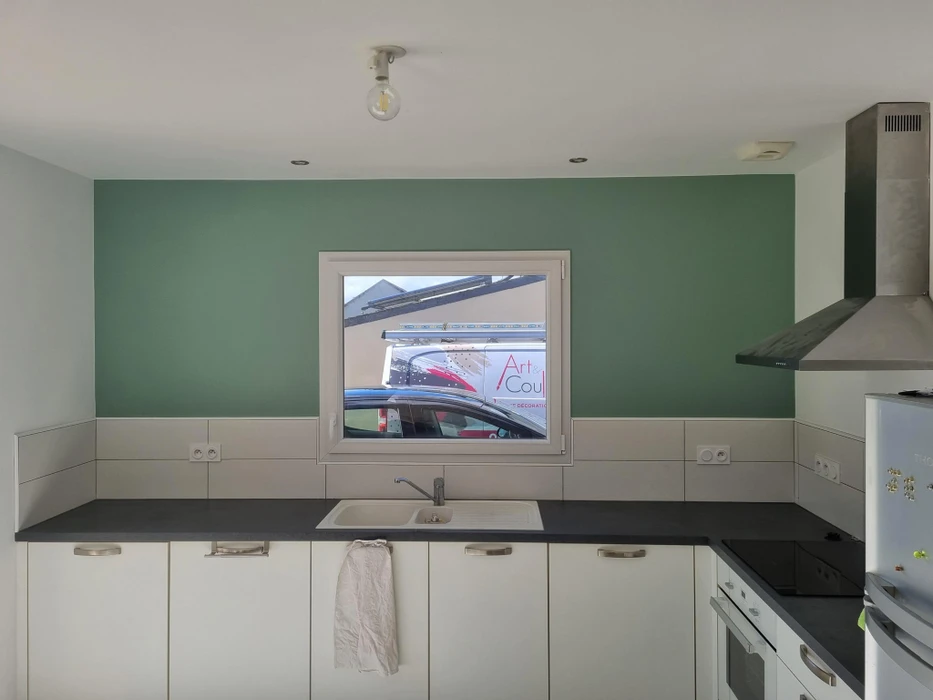 Cuisine moderne avec mur accent vert sauge, plan de travail noir, façades blanches laquées. Fenêtre fixe aluminium blanc au-dessus de l'évier encastré. Hotte aspirante inox pyramidale, carrelage métro blanc, prises électriques intégrées.