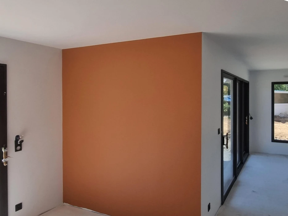 Intérieur résidentiel moderne avec mur d'accent orange terracotta contrastant avec murs blancs. Porte d'entrée sombre à gauche, grandes baies vitrées à cadre noir donnant sur l'extérieur à droite.