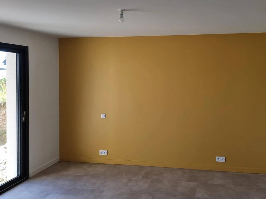 Intérieur d'une pièce en cours de finition avec mur accent peint en ocre jaune. Une personne travaille sur les finitions murales. Fenêtre avec encadrement noir sur mur blanc latéral.